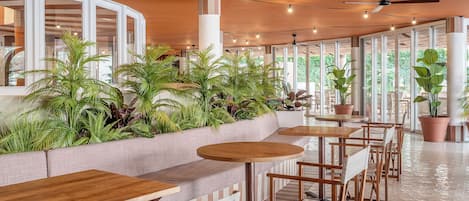 Un área de estar moderna en el interior, con mesas y sillas de madera, rodeada de amplios ventanales y plantas en macetas.