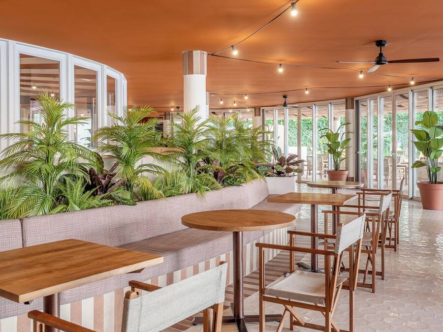 Un área de estar moderna en el interior, con mesas y sillas de madera, rodeada de amplios ventanales y plantas en macetas.