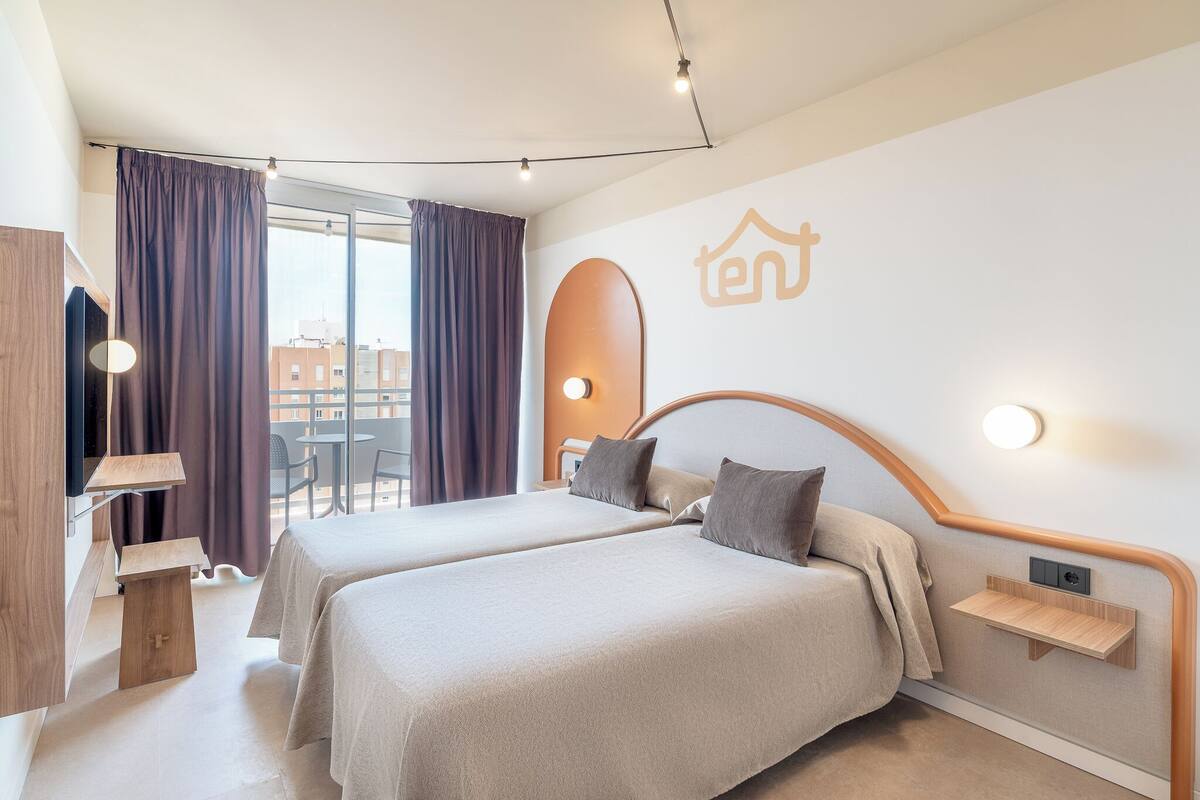 A hotel room with two beds, a TV, a desk, and a balcony with curtains.