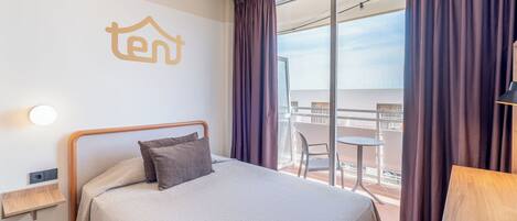 Habitación de hotel con cama, una mesita, una silla, balcón con vistas y un logotipo 'ten' en la pared.