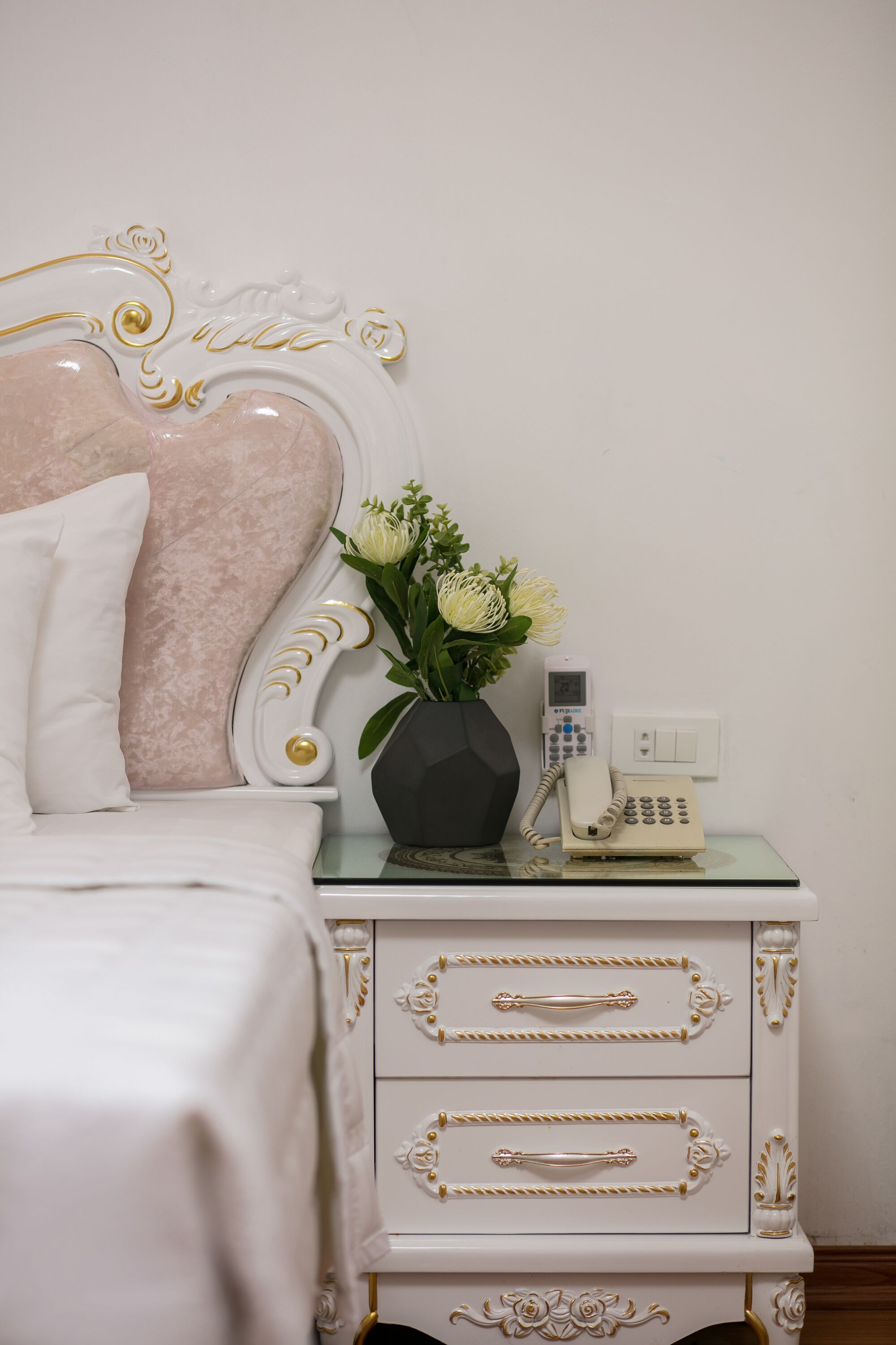 A neatly made bed with a white and pink headboard, a vase with flowers, and a telephone on a glass table beside the bed.