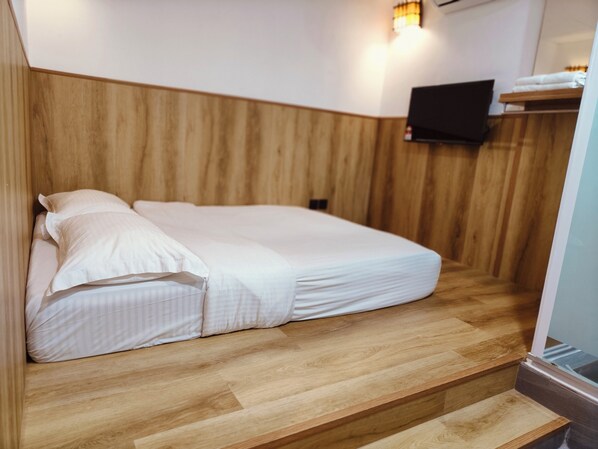 Cama com roupa branca num quarto com painéis de madeira e televisão fixada na parede.