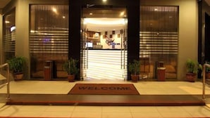 Een hotel ingang met een 'WELCOME' welkomstmat, plantenbakken en een receptiebalie zichtbaar door de glazen deuren.