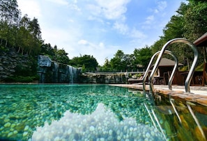 Piscina límpida com cachoeira ao fundo, cadeiras de descanso e deck de madeira.