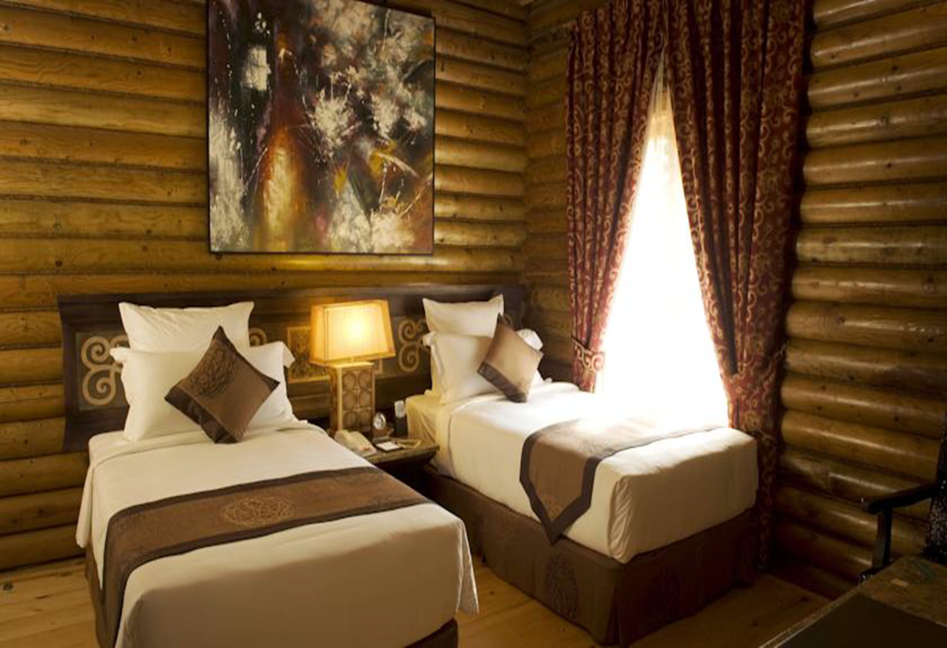 A log cabin room with two beds, a painting on the wall, and a window with curtains.
