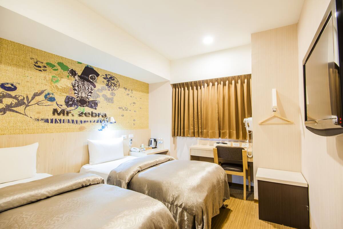 A hotel room with two beds, a desk, a chair, a TV, and a wall with a zebra mural.