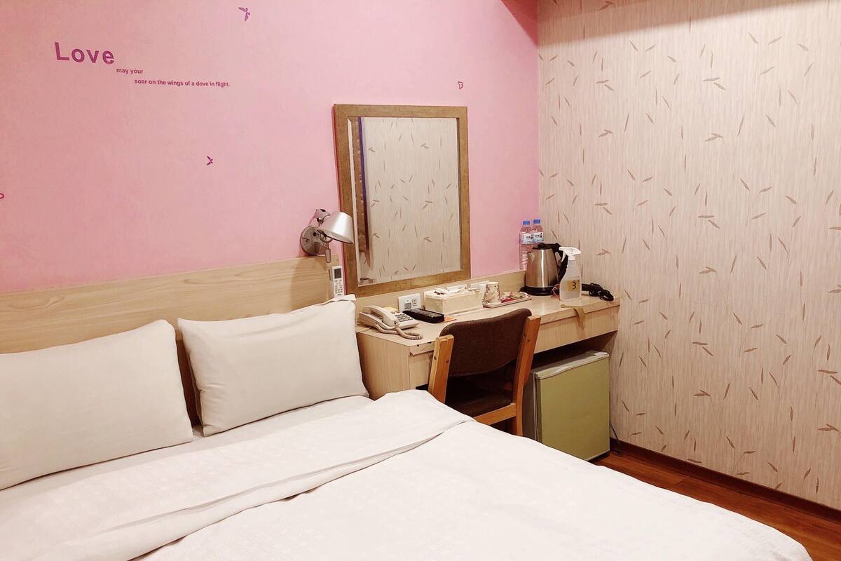 A hotel room with a bed, a desk, a chair, a small fridge, a mirror, and a pink wall with a 'Love' sign.