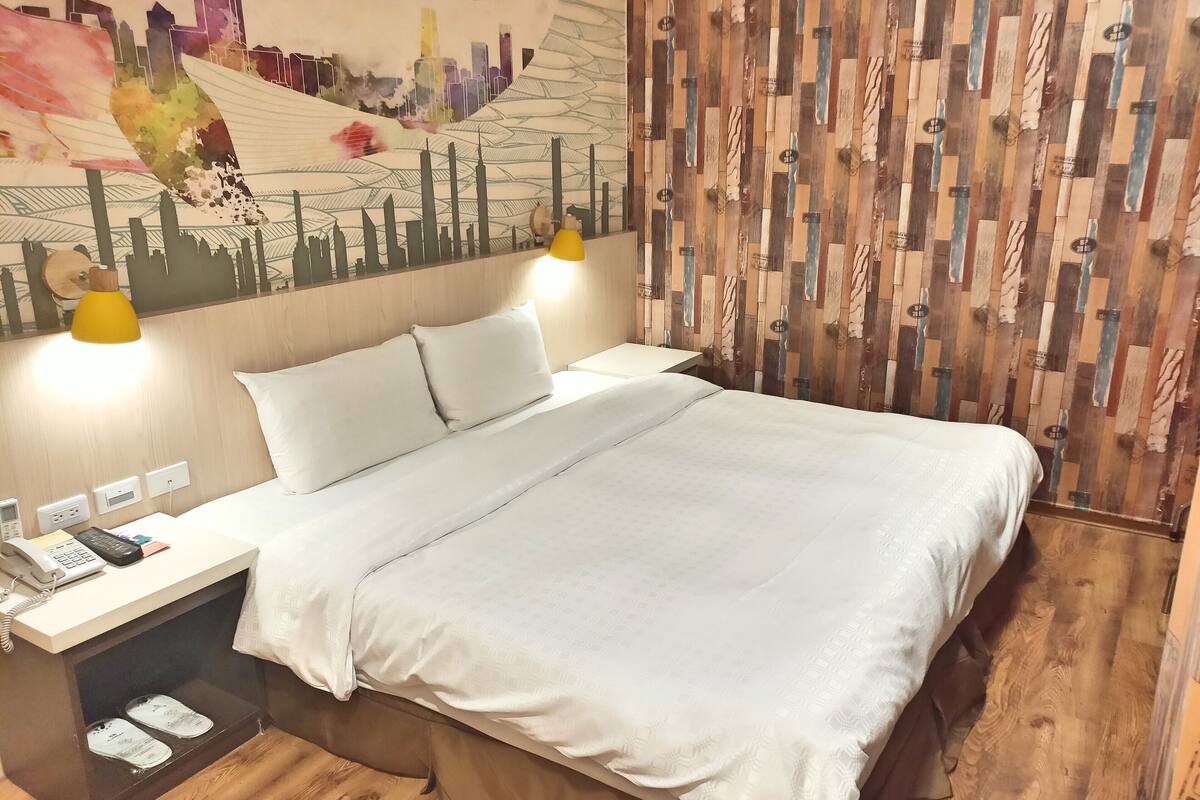 A hotel room with a bed, bedside lamps, a phone, and a wall with a cityscape mural.