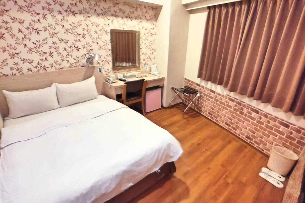 A hotel room with a bed, a desk, a mirror, a window with curtains, and a brick wall.