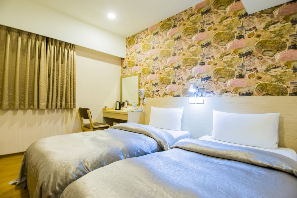 A hotel room with two beds, a stone-patterned wall, a desk with a chair, and a window with curtains.