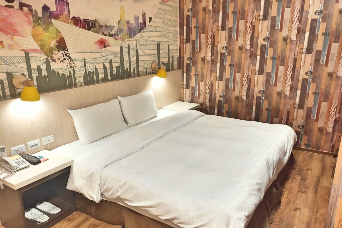 A hotel room with a bed, bedside table, lamp, and a wall with a cityscape mural.