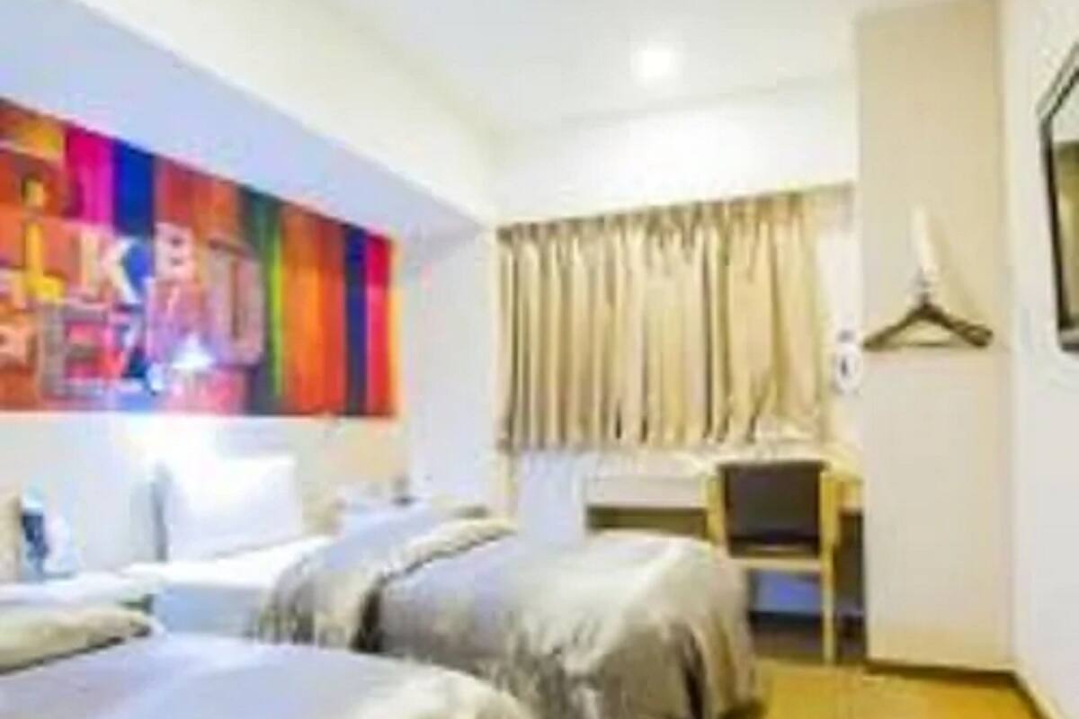 A hotel room with two beds, a desk, a chair, a colorful artwork on the wall, and a window with curtains.