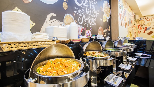 A buffet setup with various food items in large metal containers, a cake on a table, and a wall with a coffee-themed mural. - Sanduo Hotel (Kaohsiung)