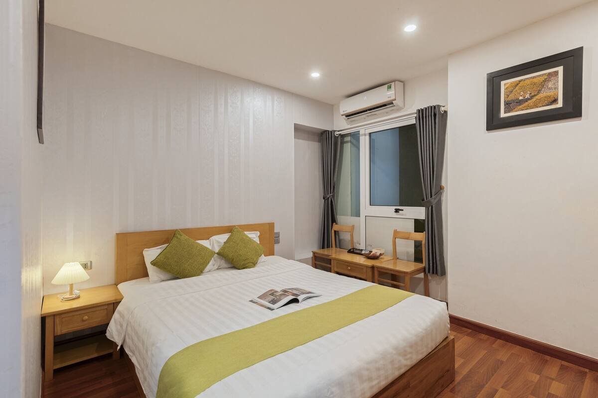 Une chambre d’hôtel comprenant un lit, une table de chevet avec une lampe, un bureau avec des chaises, une fenêtre avec des rideaux et un appareil de climatisation.