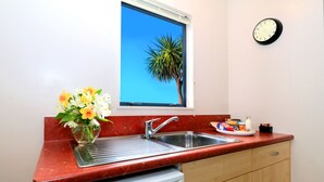 A kitchen with a red countertop, a sink, a dish rack, a white dishwasher, a window with a view of a palm tree, and a wall clock. - Bella Vista Motel Invercargill (Invercargill)