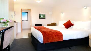 A hotel room with a large bed, a desk with a chair, a small table with flowers, and a door leading to a bathroom.