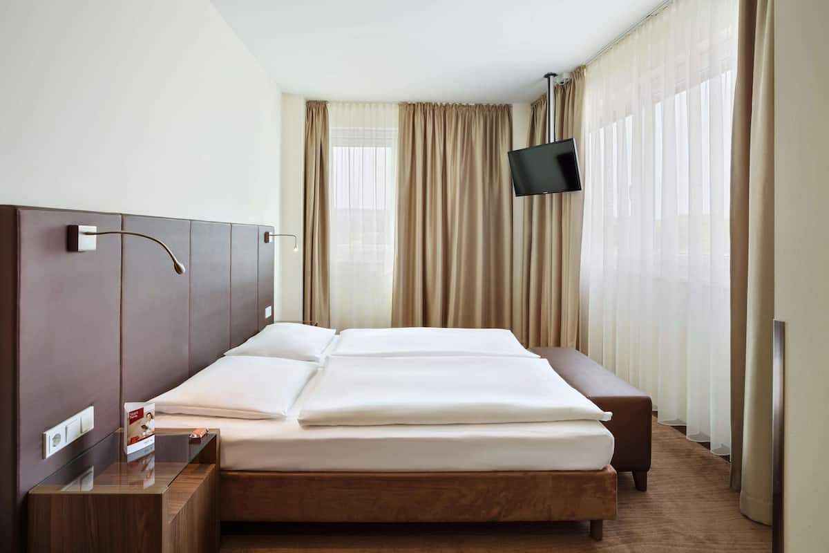 Una habitación de hotel moderna con una cama grande, una mesita de noche, un televisor de pantalla plana y una ventana con cortinas.