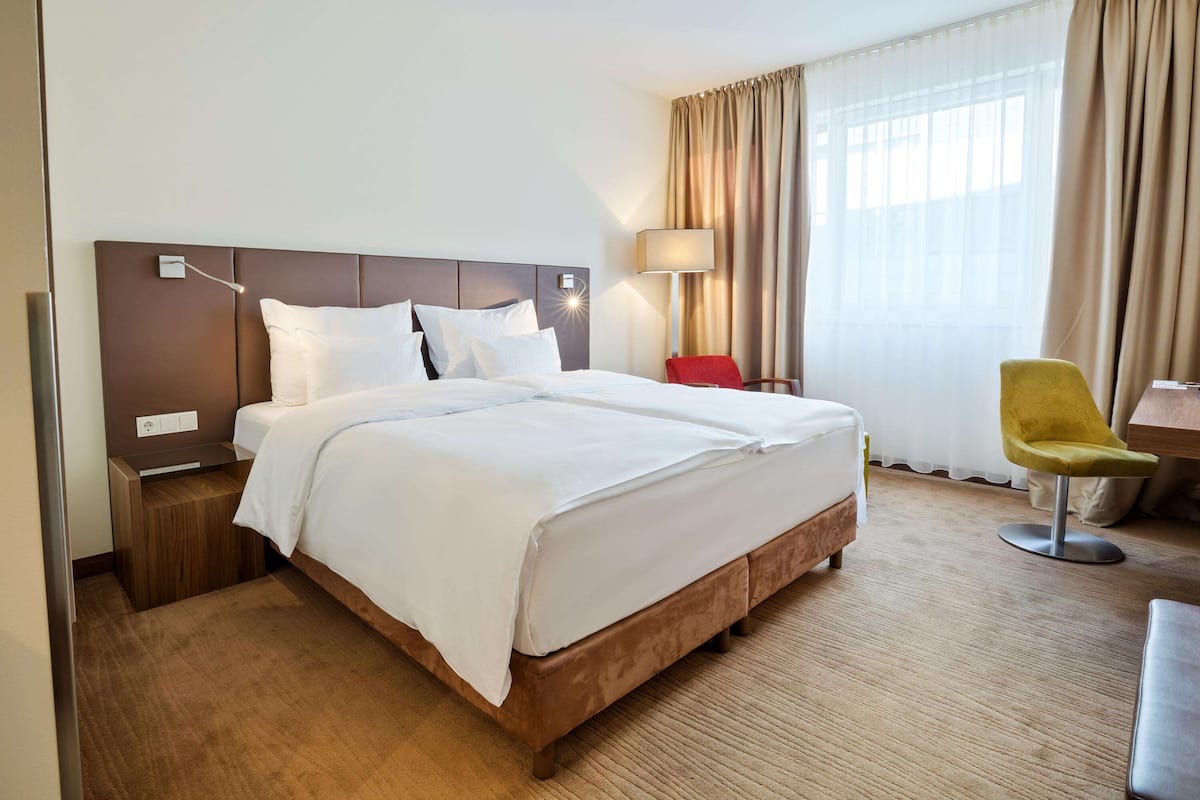 Una habitación de hotel con una cama grande, una mesita de noche, un escritorio, una silla y un ventanal con cortinas.