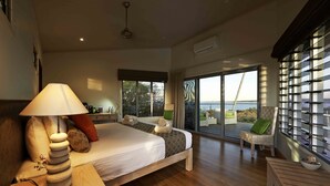 A bedroom with a large bed, a bedside table with a lamp, a chair by the window, and a view of the sea. - The Berkeley River Lodge (Oombulgurri)