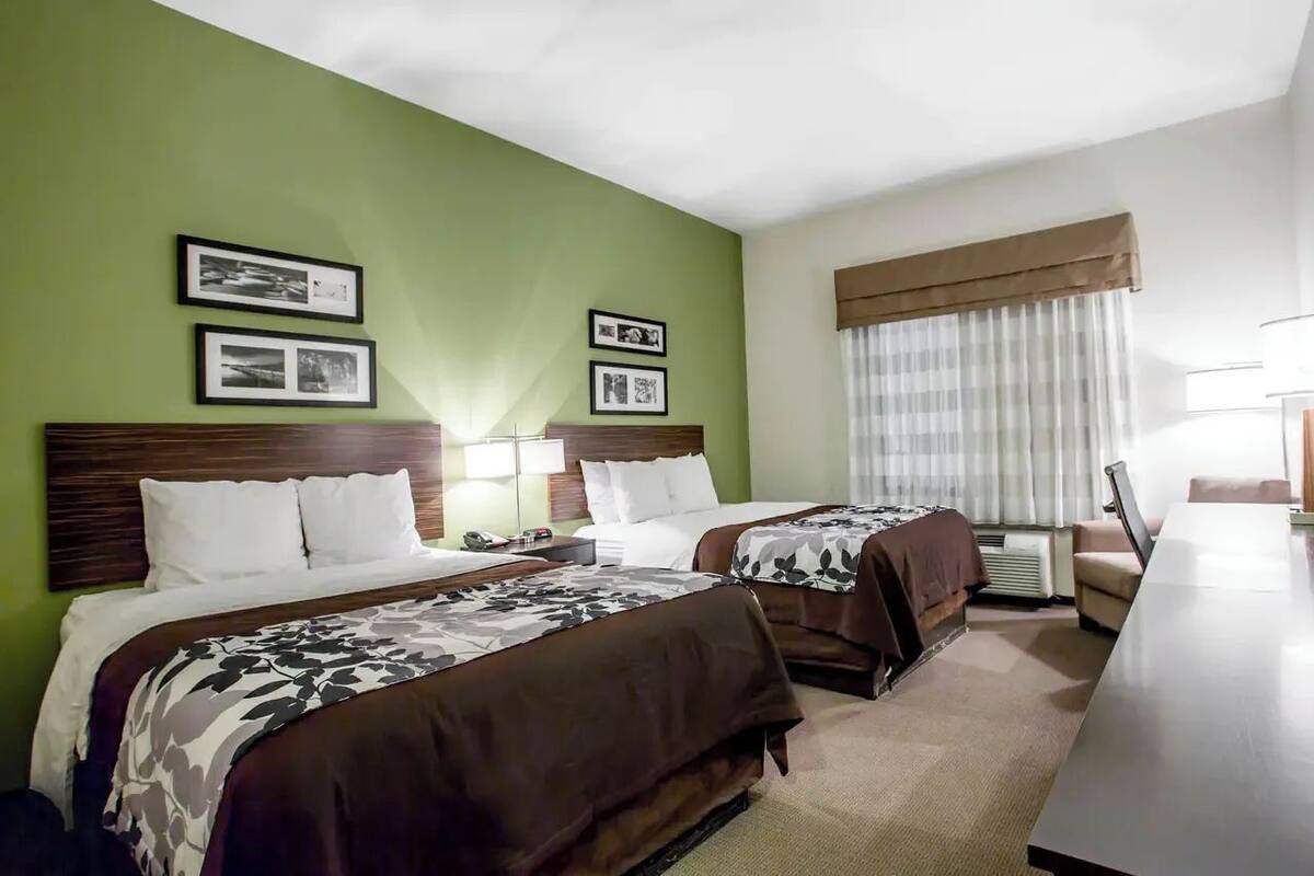 A hotel room with two beds, a desk, a chair, and a large window with curtains.