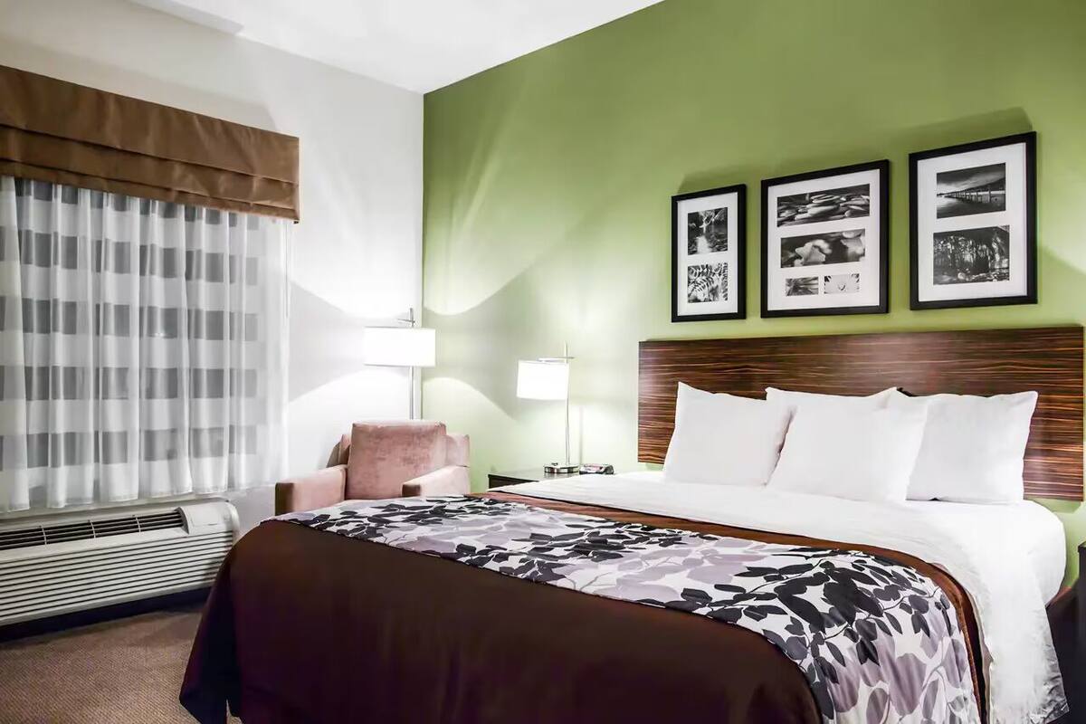 A hotel room with a bed, a chair, a bedside table, a lamp, and framed pictures on the wall.