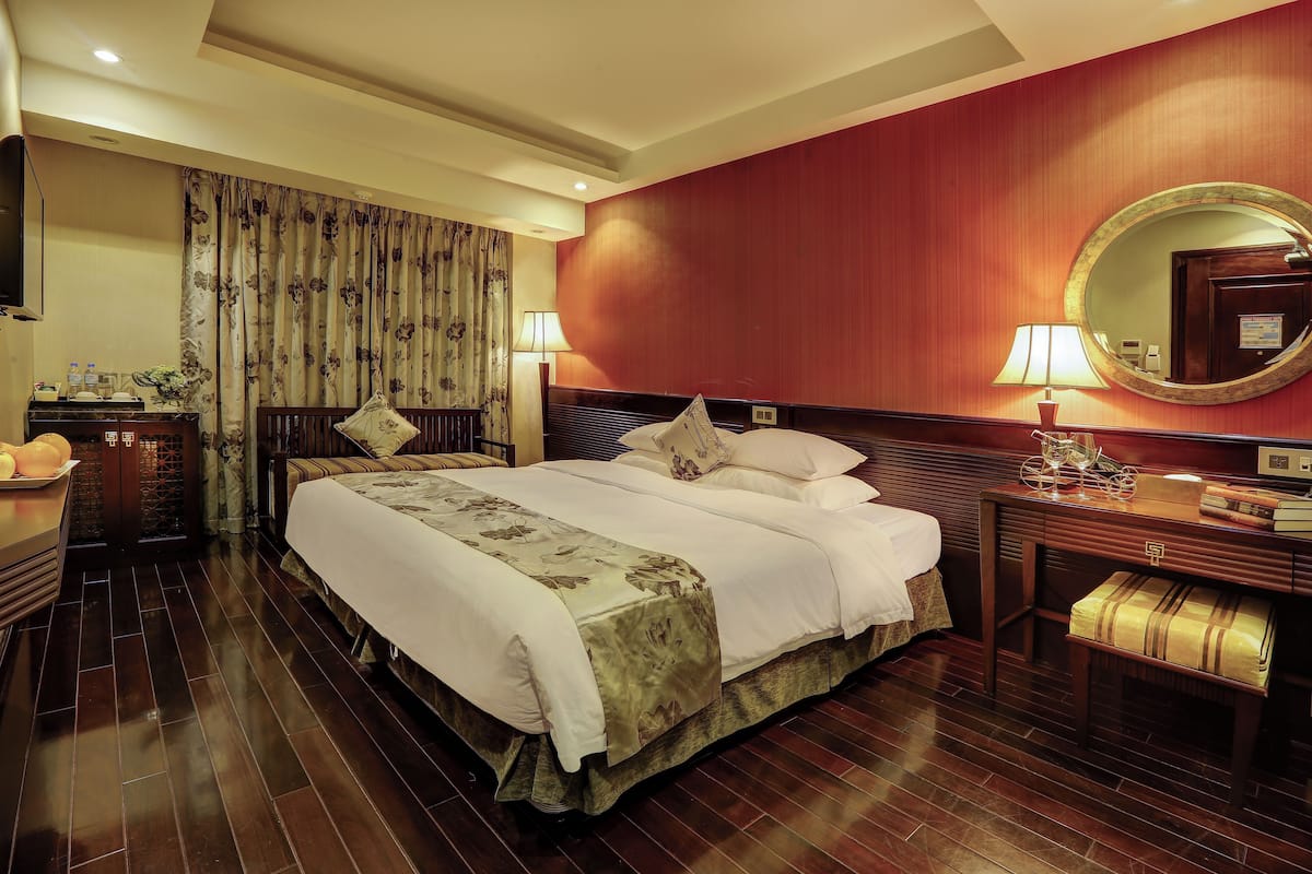 Une chambre d’hôtel avec un grand lit, un plancher en bois, un bureau avec une lampe, un miroir et une fenêtre avec des rideaux.