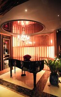 A grand piano in a well-lit lobby with a chandelier, a potted plant, and a vase with red flowers.