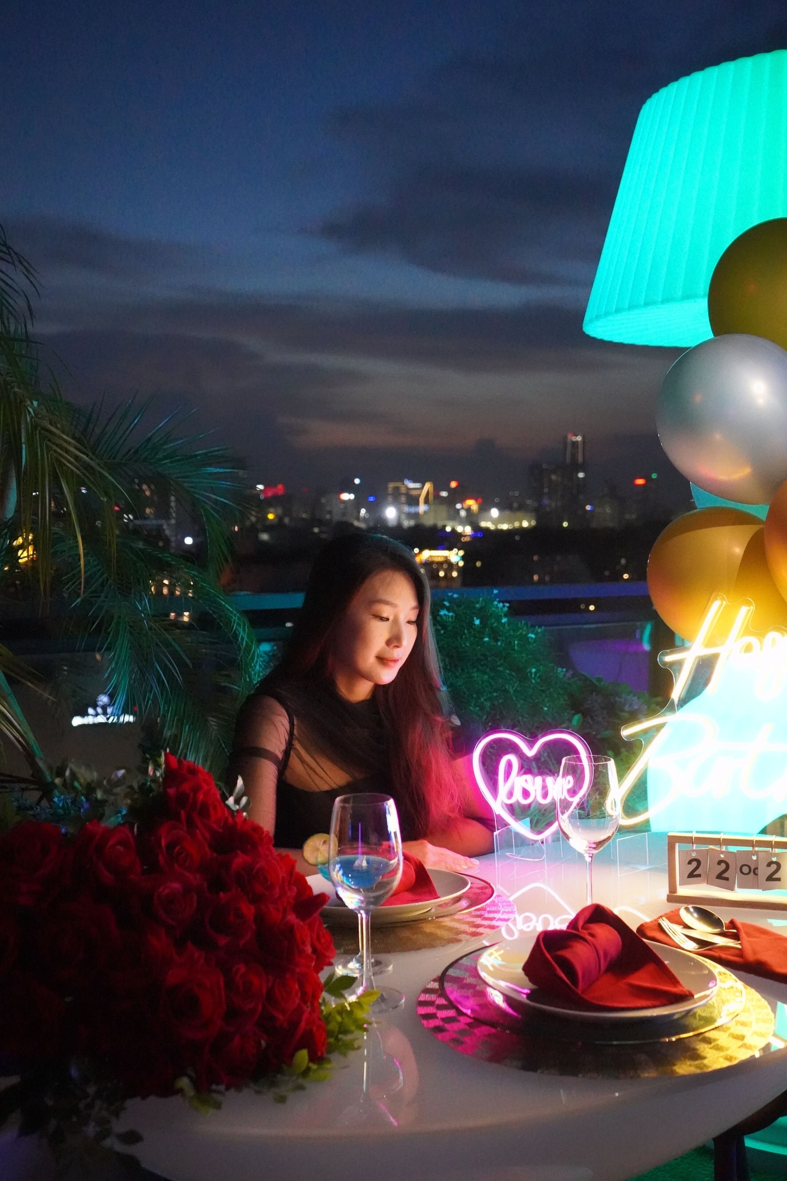 Une table dressée pour deux, avec un panneau lumineux au néon, un bouquet de roses rouges et, en arrière-plan, une vue sur le paysage urbain.