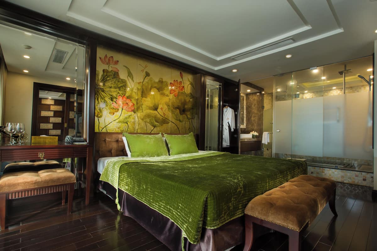 Une chambre spacieuse avec un grand lit, un banc, une coiffeuse avec un miroir et une salle de bain dotée d’une douche à parois de verre.