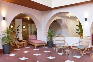 Un espacio interior amplio con una cama, sillas de mimbre, plantas en macetas y piso de baldosas.