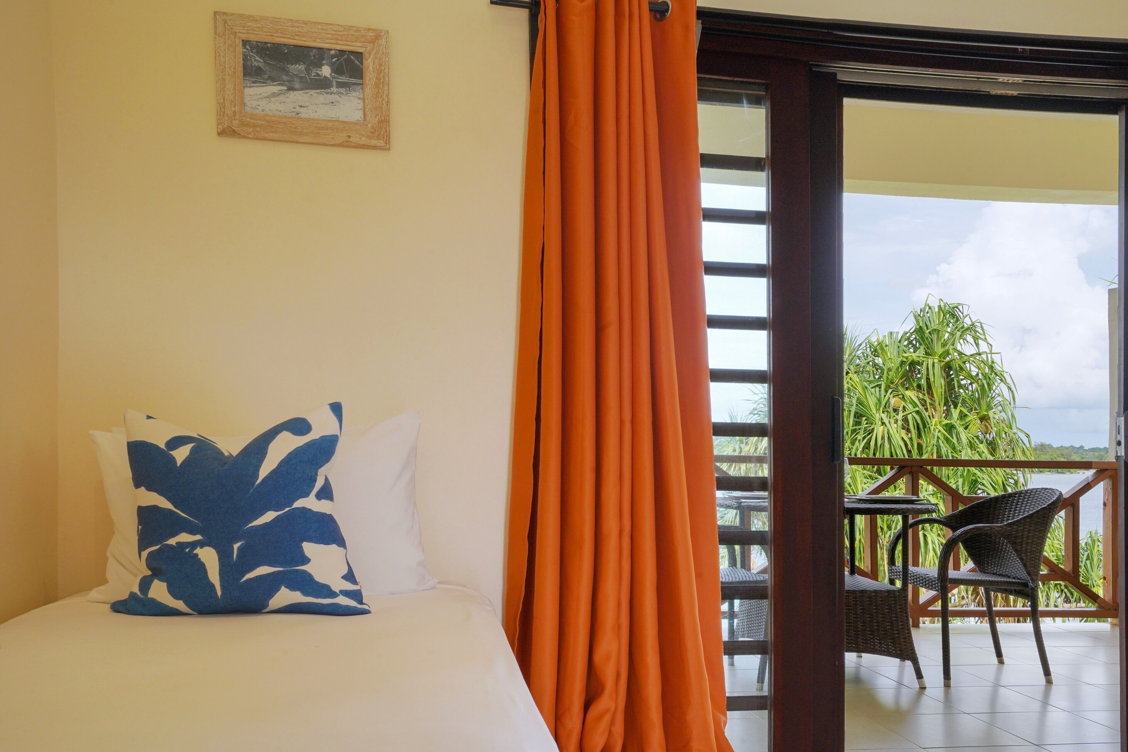 Une pièce avec un lit, un grand rideau orange, un tableau encadré au mur et une vue sur un balcon avec des chaises et de la verdure.