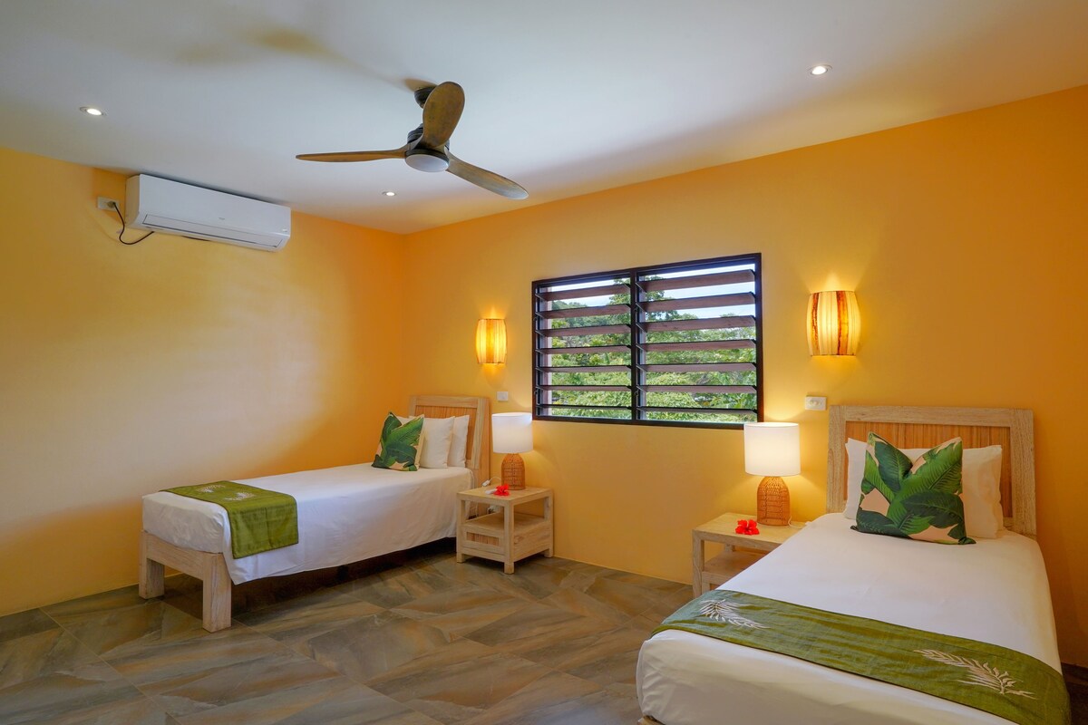 A hotel room with two single beds, a ceiling fan, an air conditioning unit, a window with blinds, and two bedside lamps.