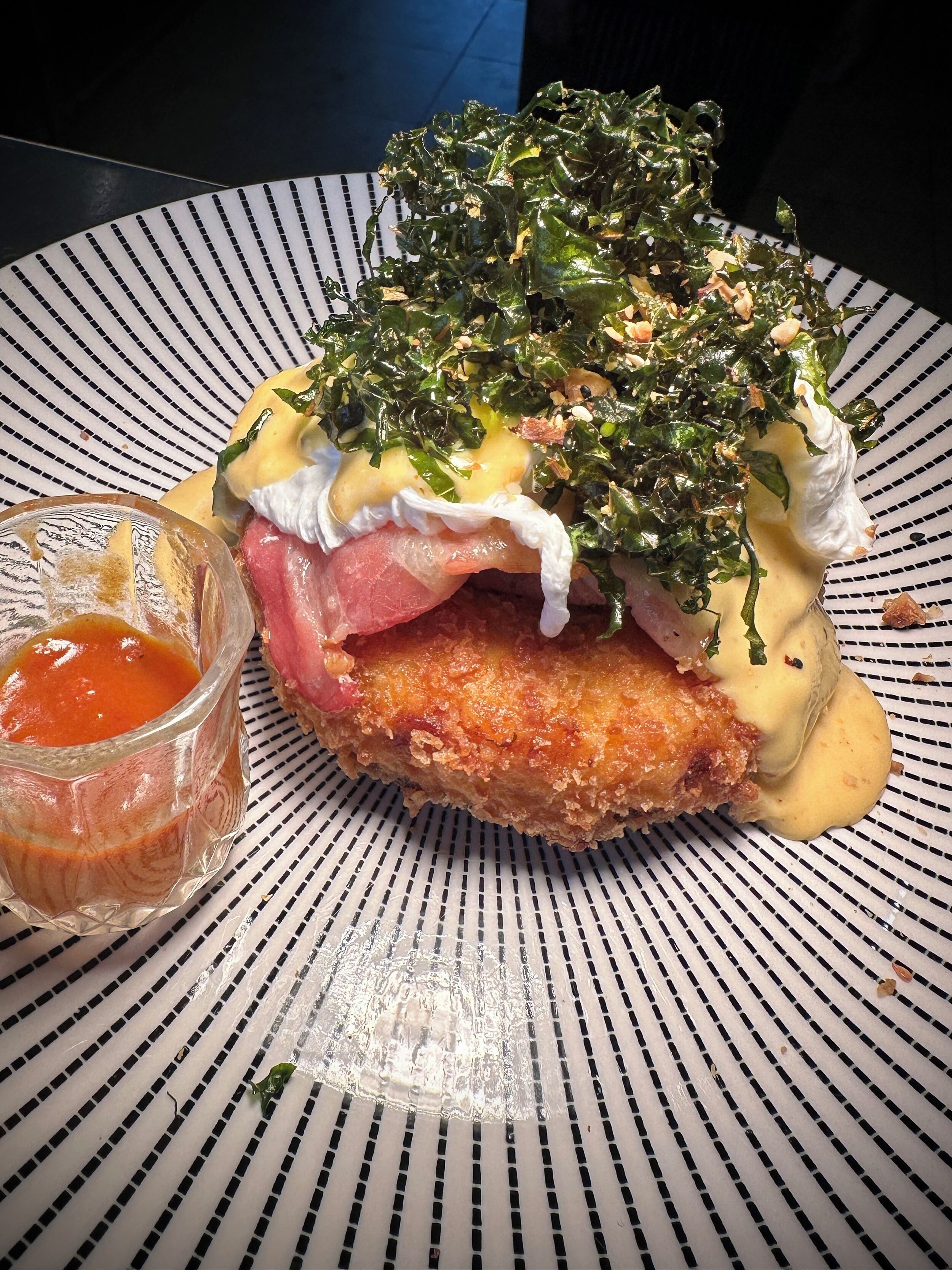 Een bord met een gepaneerde schnitzel, groenten, saus en een klein glaasje saus.