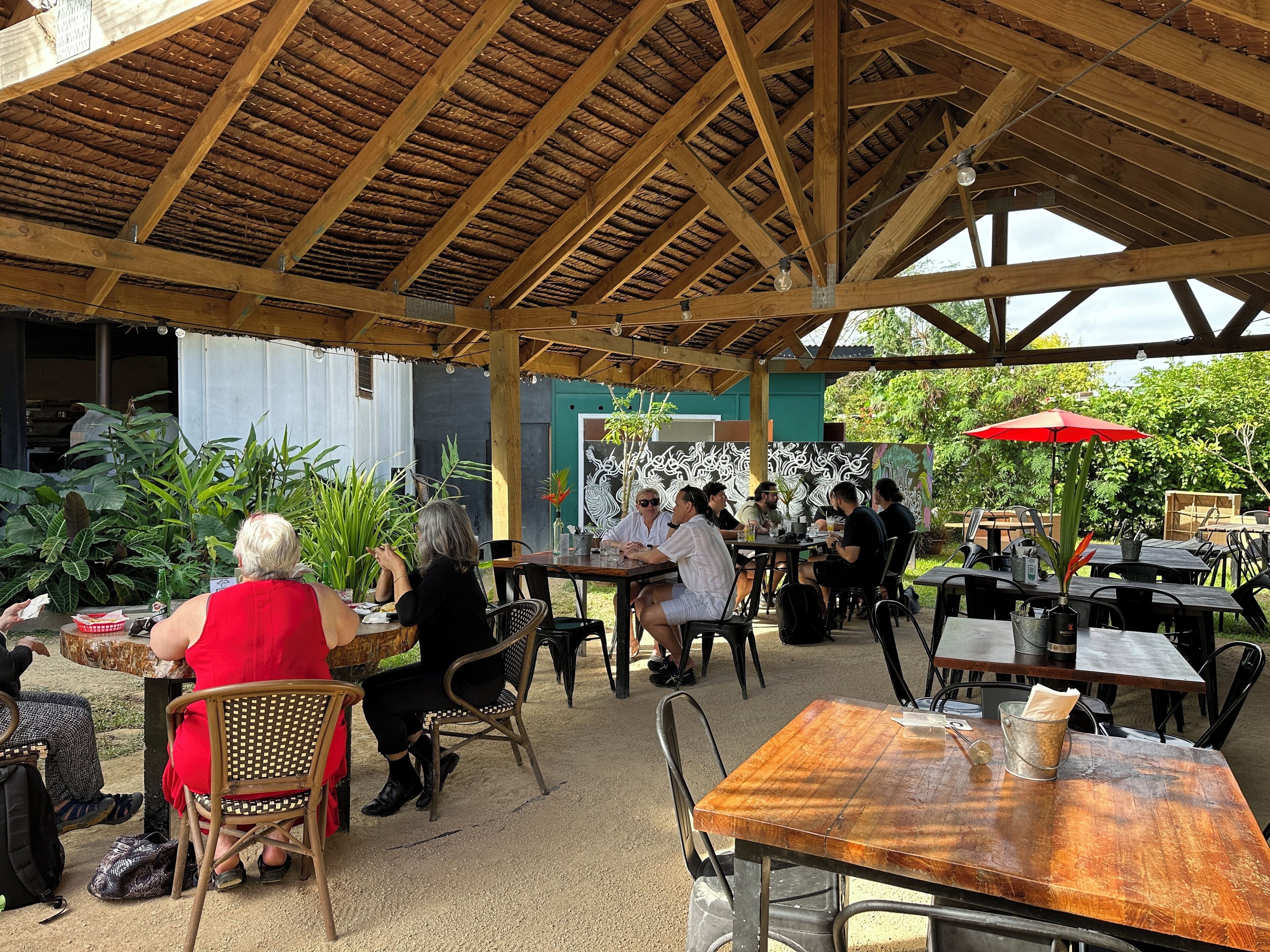 An outdoor patio area with wooden tables and chairs, surrounded by greenery and a thatched roof structure.