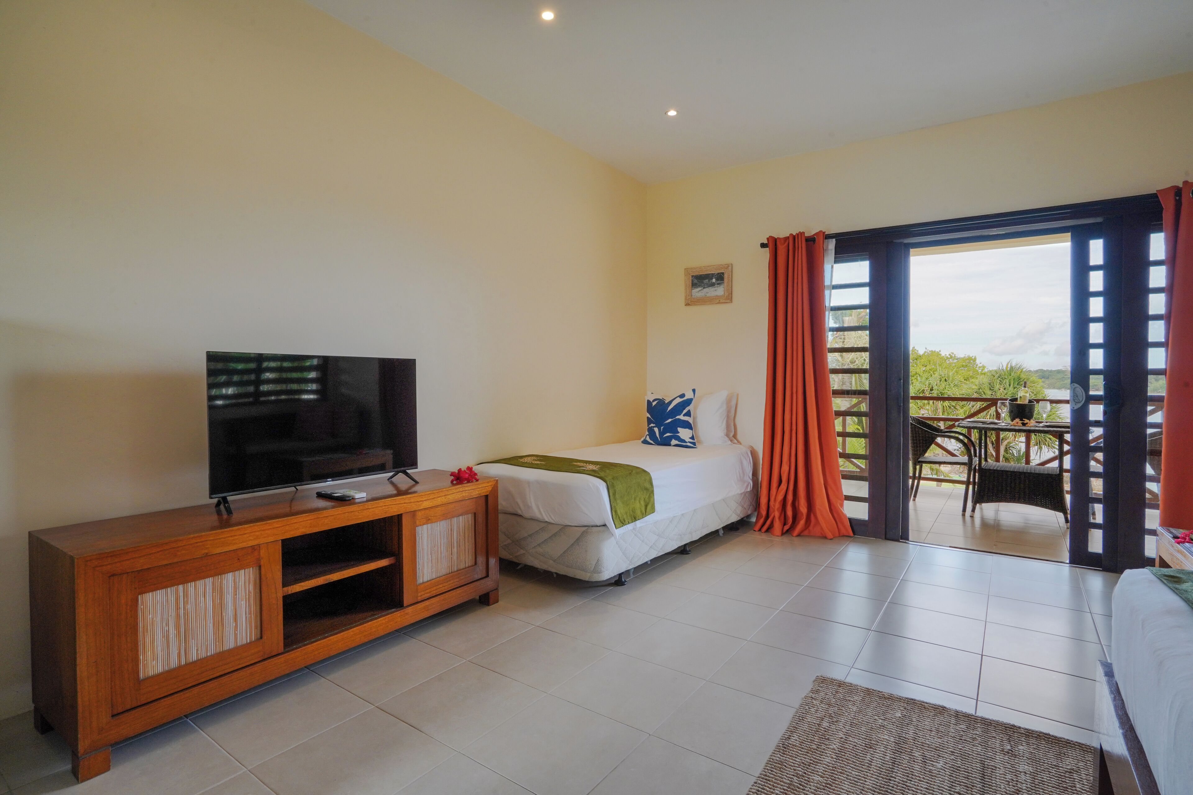 A bedroom with a bed, a TV on a wooden cabinet, a balcony with a view, and a small table with chairs.