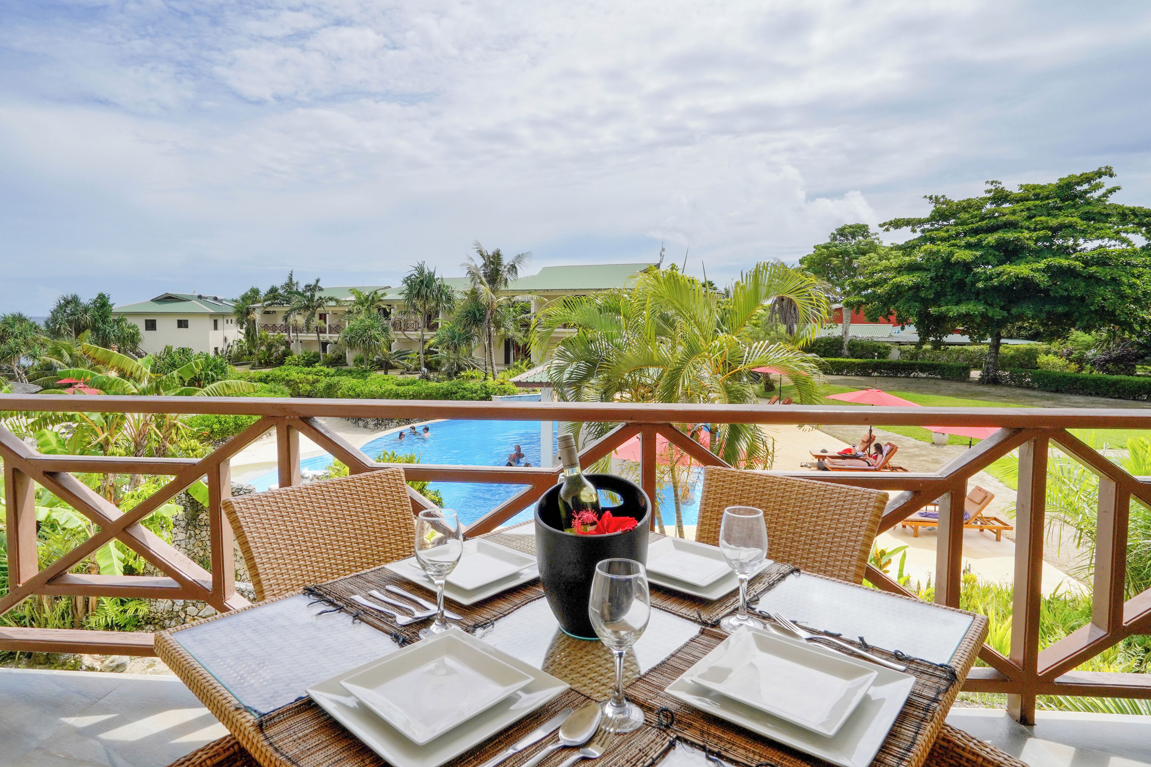 A dining table set with white plates, cutlery, and wine glasses, overlooking a pool and a tropical landscape.