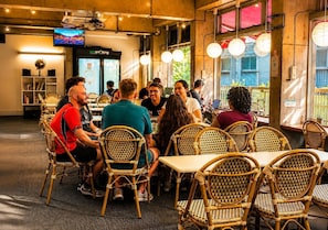 A group of people seated at tables in a room with wicker chairs, a television on the wall, and a window with a view of greenery. - Sakura Hostel Asakusa (Tokyo)