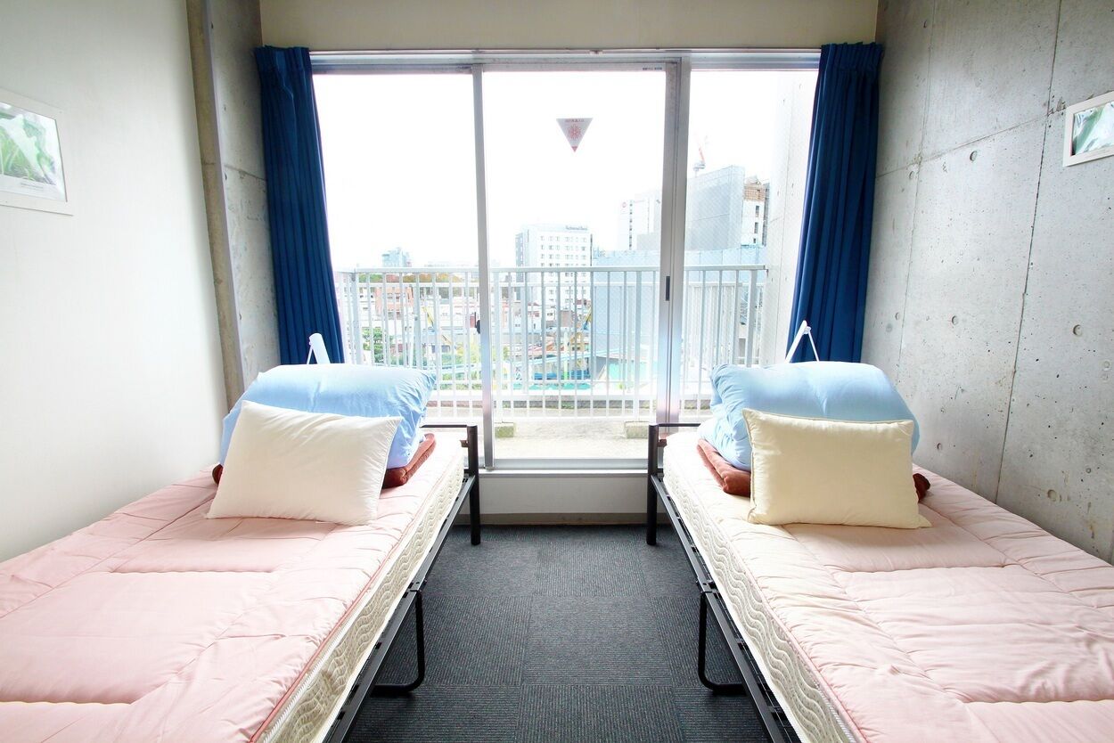 A room with two beds, a window with curtains, and a view of buildings outside.