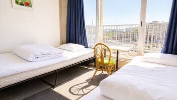A hotel room with two single beds, a chair, a window with a view of buildings, and a framed picture on the wall.