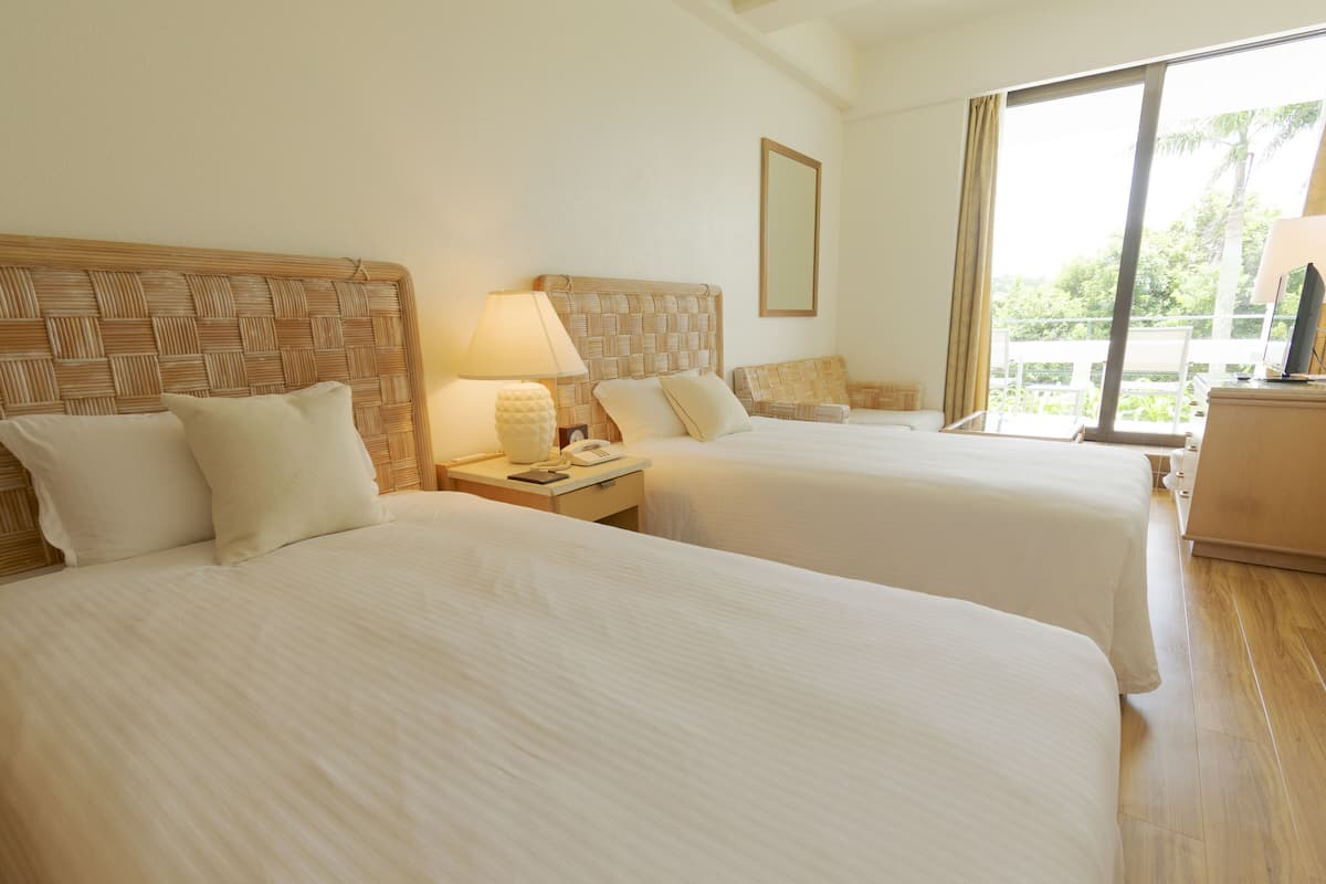 A hotel room with two beds, a nightstand with a lamp, a telephone, and a window with a view of greenery.