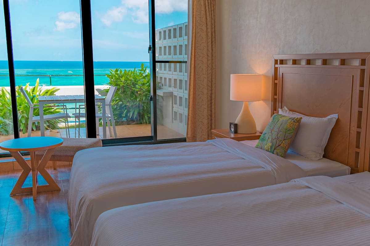 A hotel room with two beds, a balcony with a view of the ocean, a small table, and a lamp.