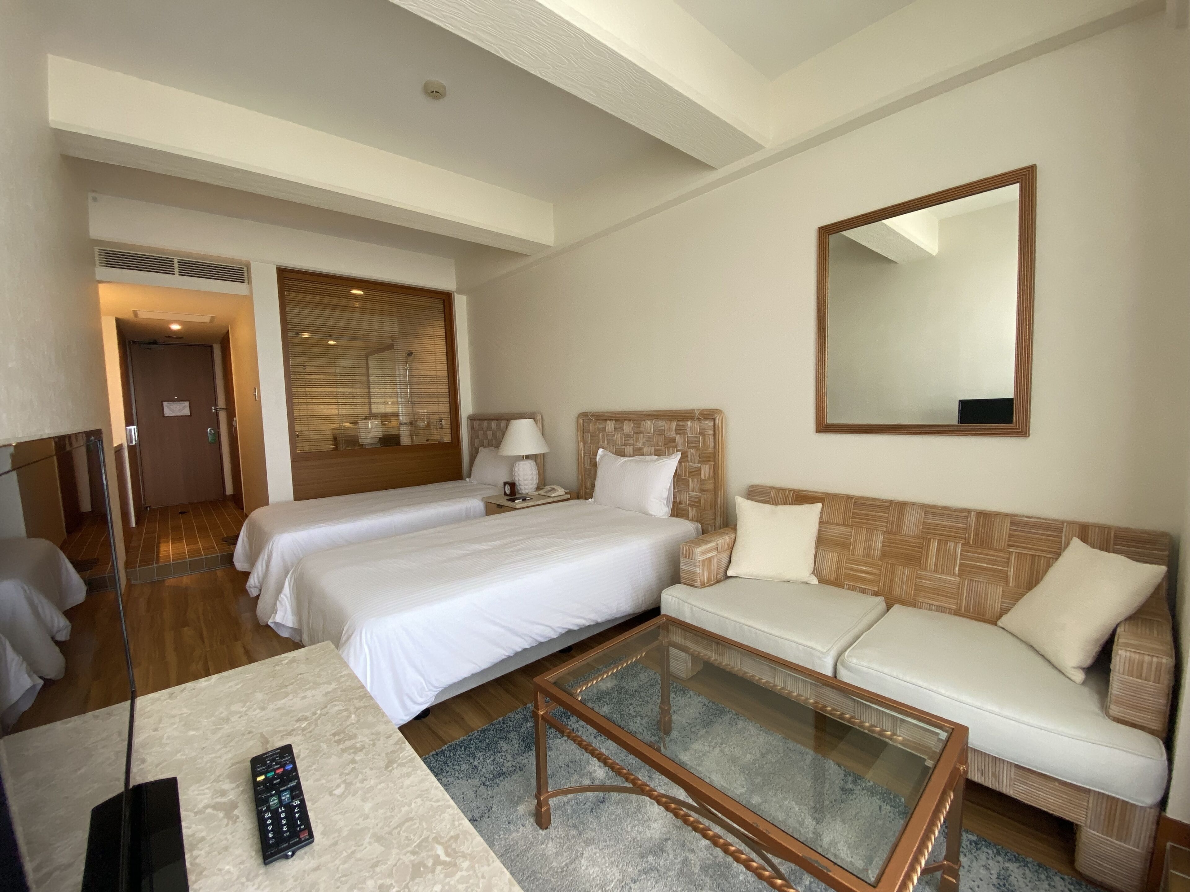 Ein Hotelzimmer mit zwei Betten, einem Sofa, einem Couchtisch, einem Spiegel und einem Fernseher.