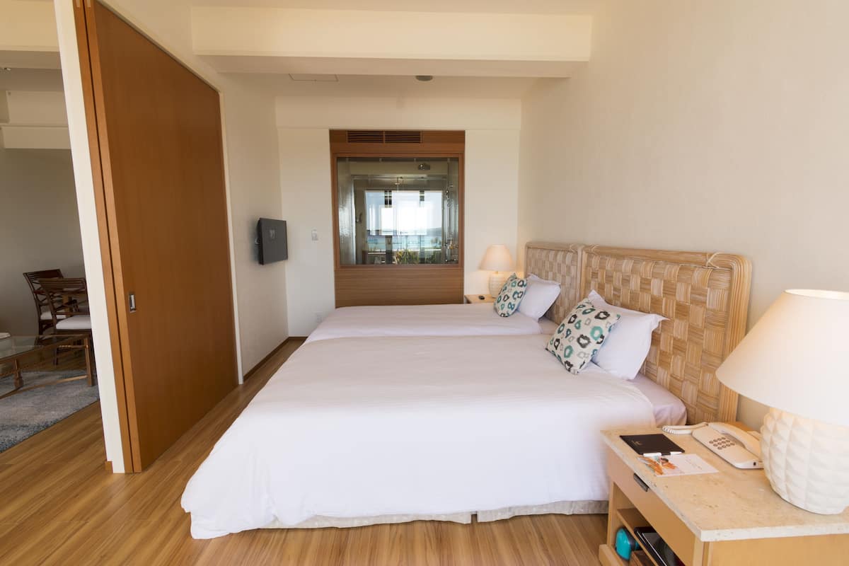 A hotel room with a large bed, a bedside table with a lamp, and a view of the dining area through a glass door.