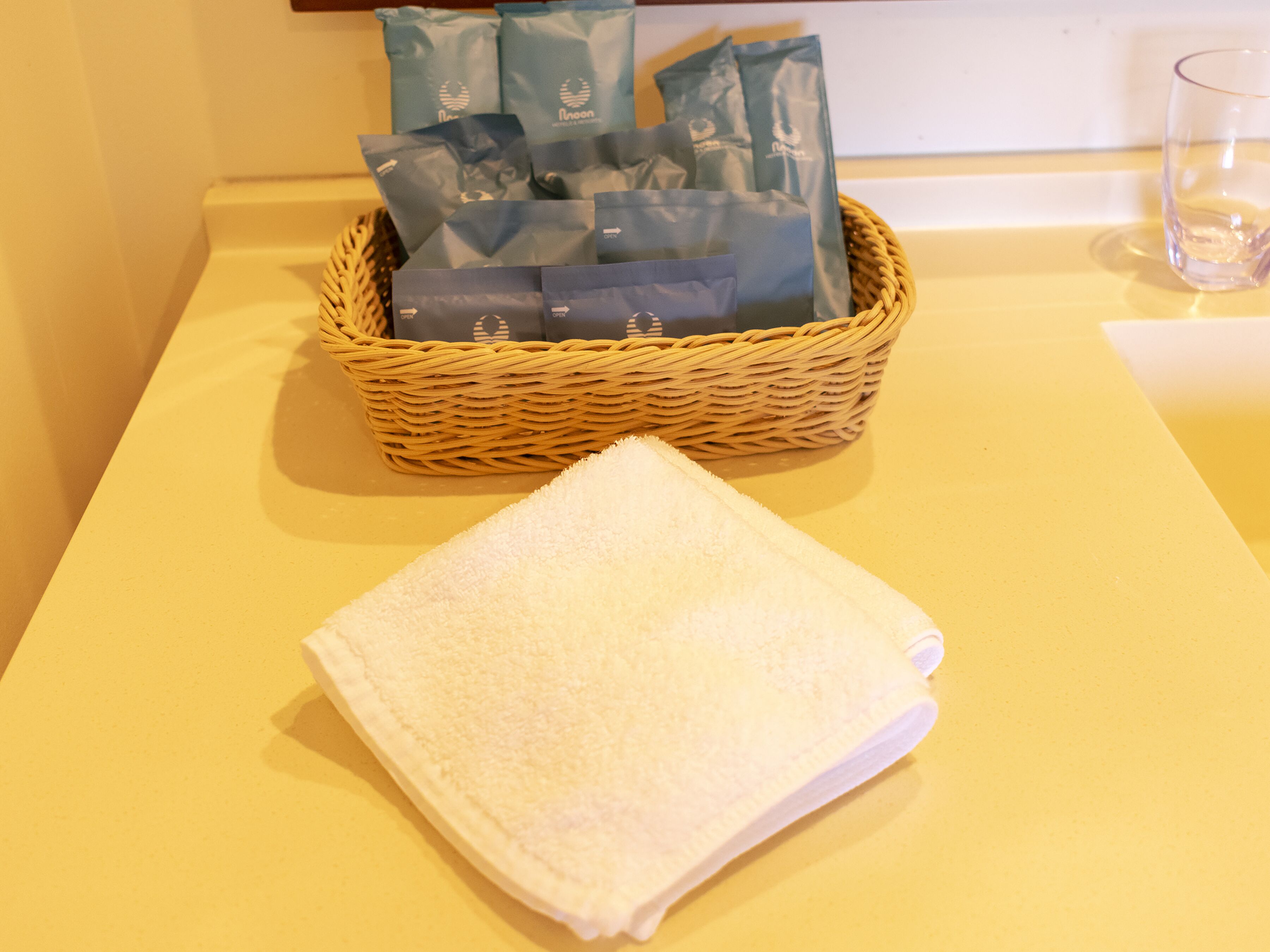 Un panier en osier contenant plusieurs paquets bleus, posé sur une table jaune, à côté d’une serviette blanche pliée.