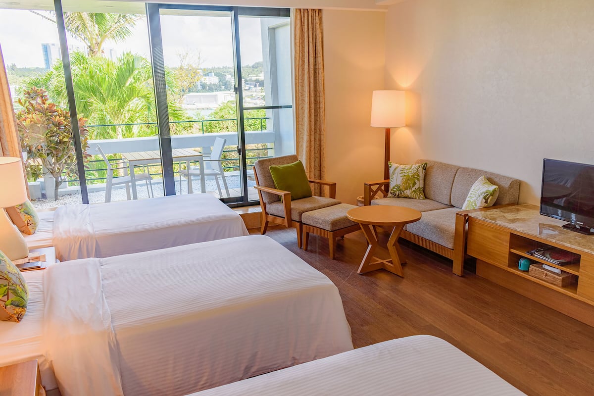 A hotel room with two beds, a sofa, a small table, a TV, and a balcony with a view of greenery and buildings.