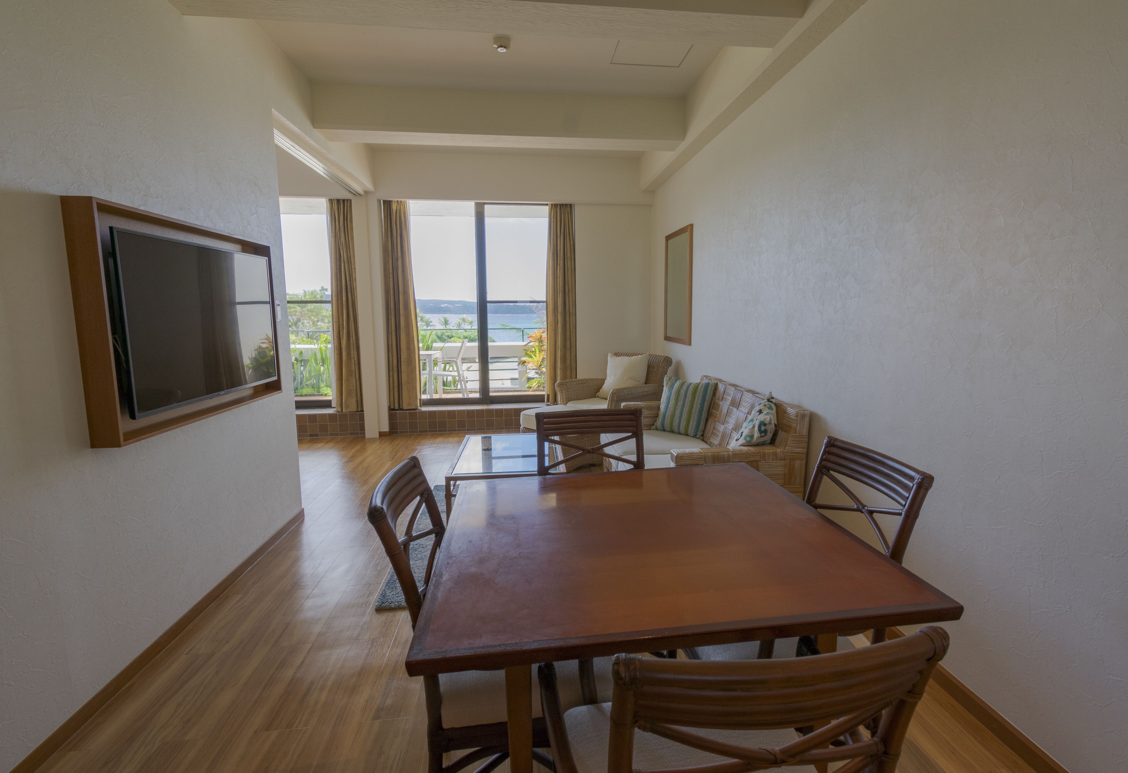 A spacious living room with a wooden dining table, comfortable seating, a flat-screen TV, and large windows offering a view of the outdoors.
