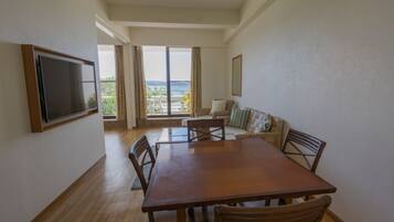 A spacious living room with a wooden dining table, comfortable seating, a flat-screen TV, and large windows offering a view of the outdoors.