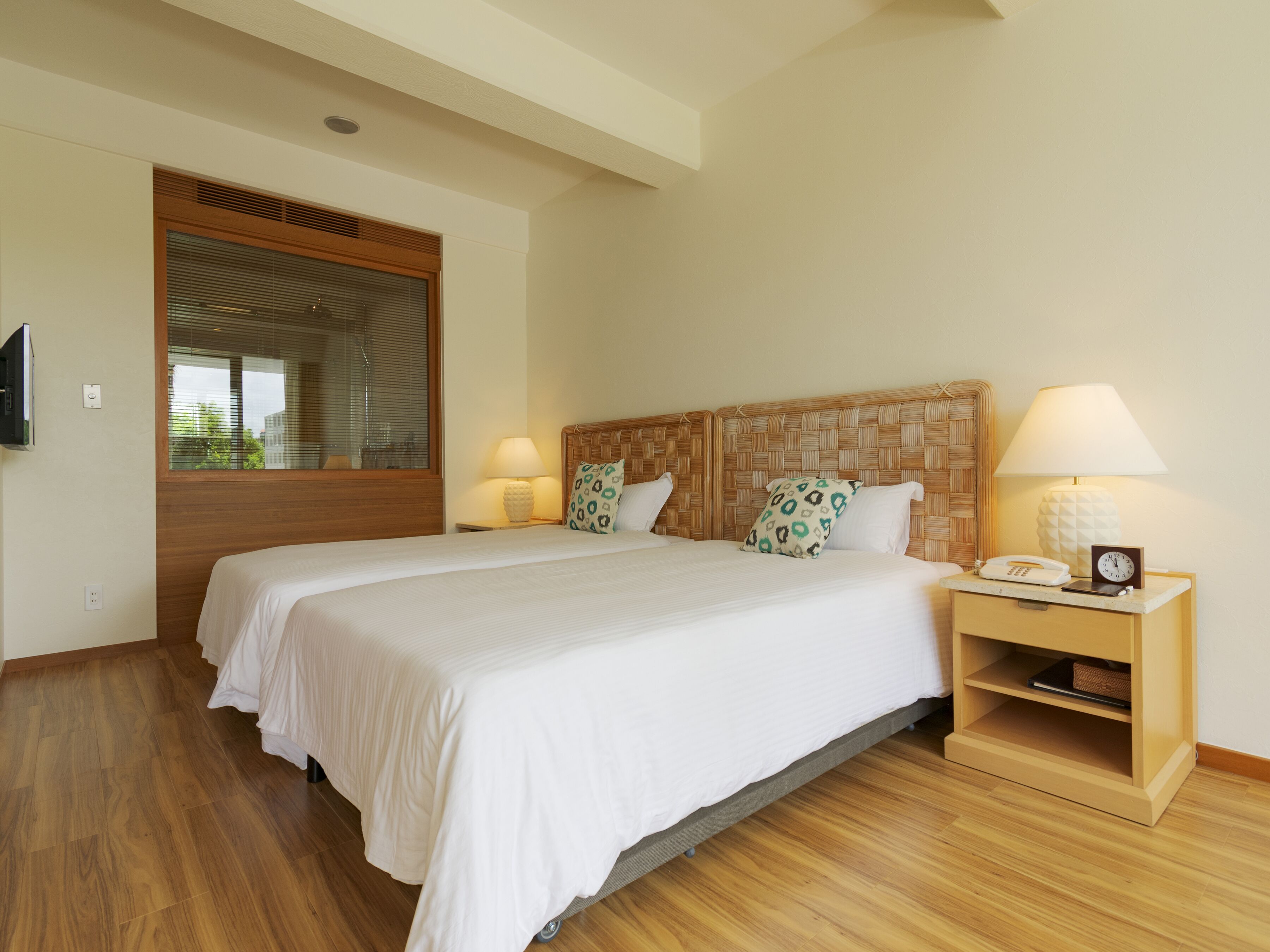 A double bed with a woven headboard, two bedside lamps, a bedside telephone, and a small shelf with a book and a decorative item.