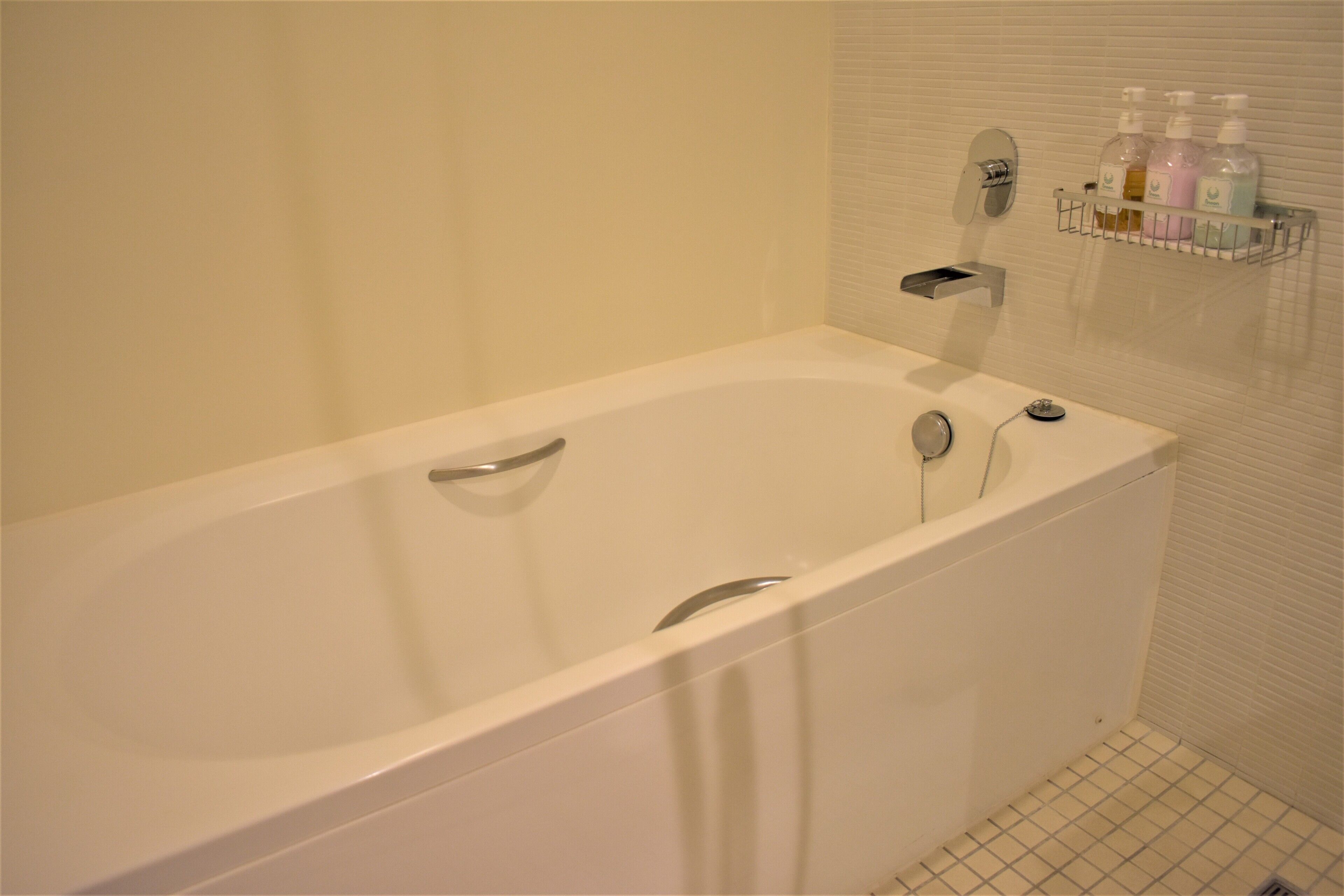 A modern bathroom with a white bathtub, a handheld showerhead, and a soap dispenser shelf.