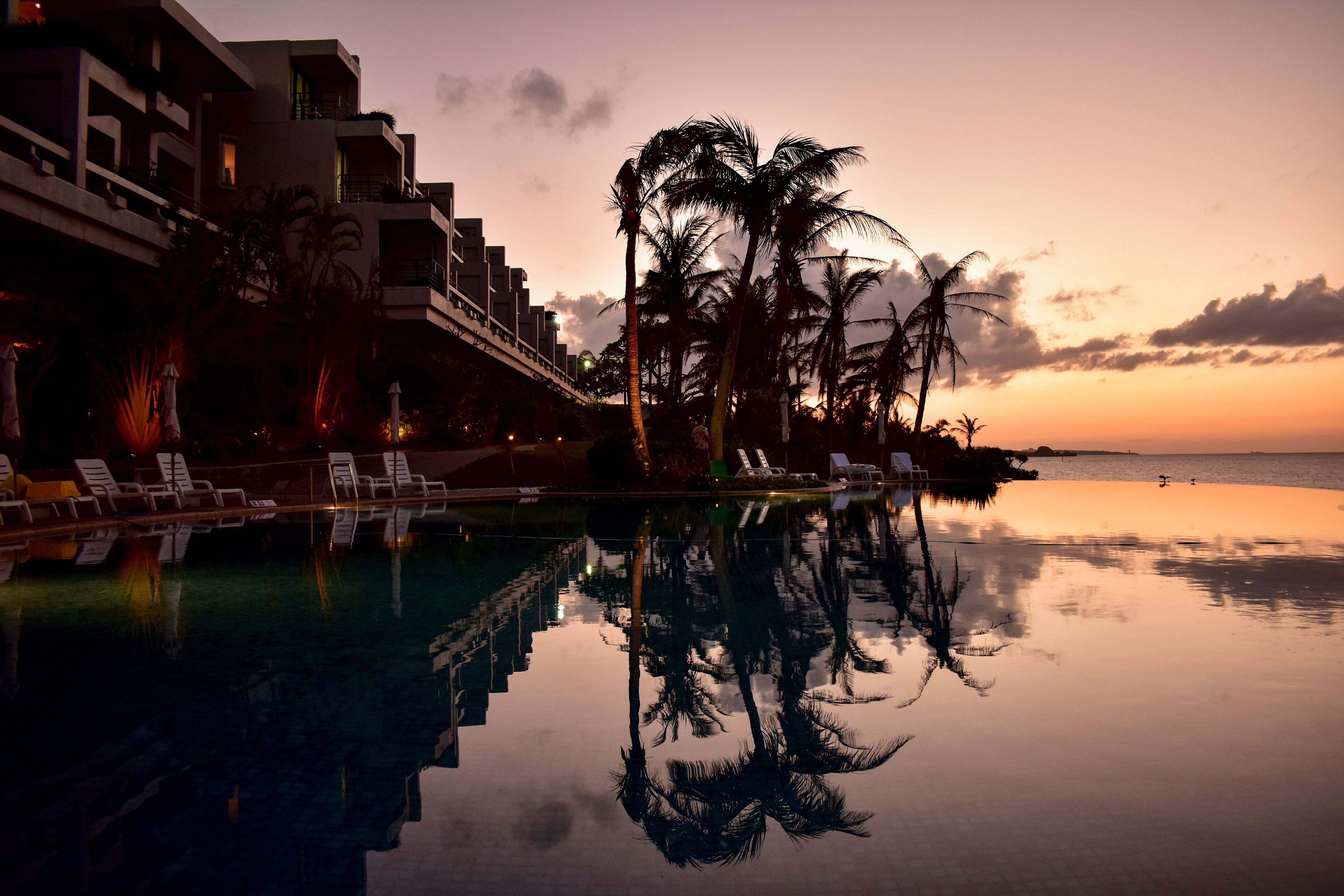 Une vue sur la piscine au coucher du soleil, avec le reflet des palmiers et d’un bâtiment dans l’eau.